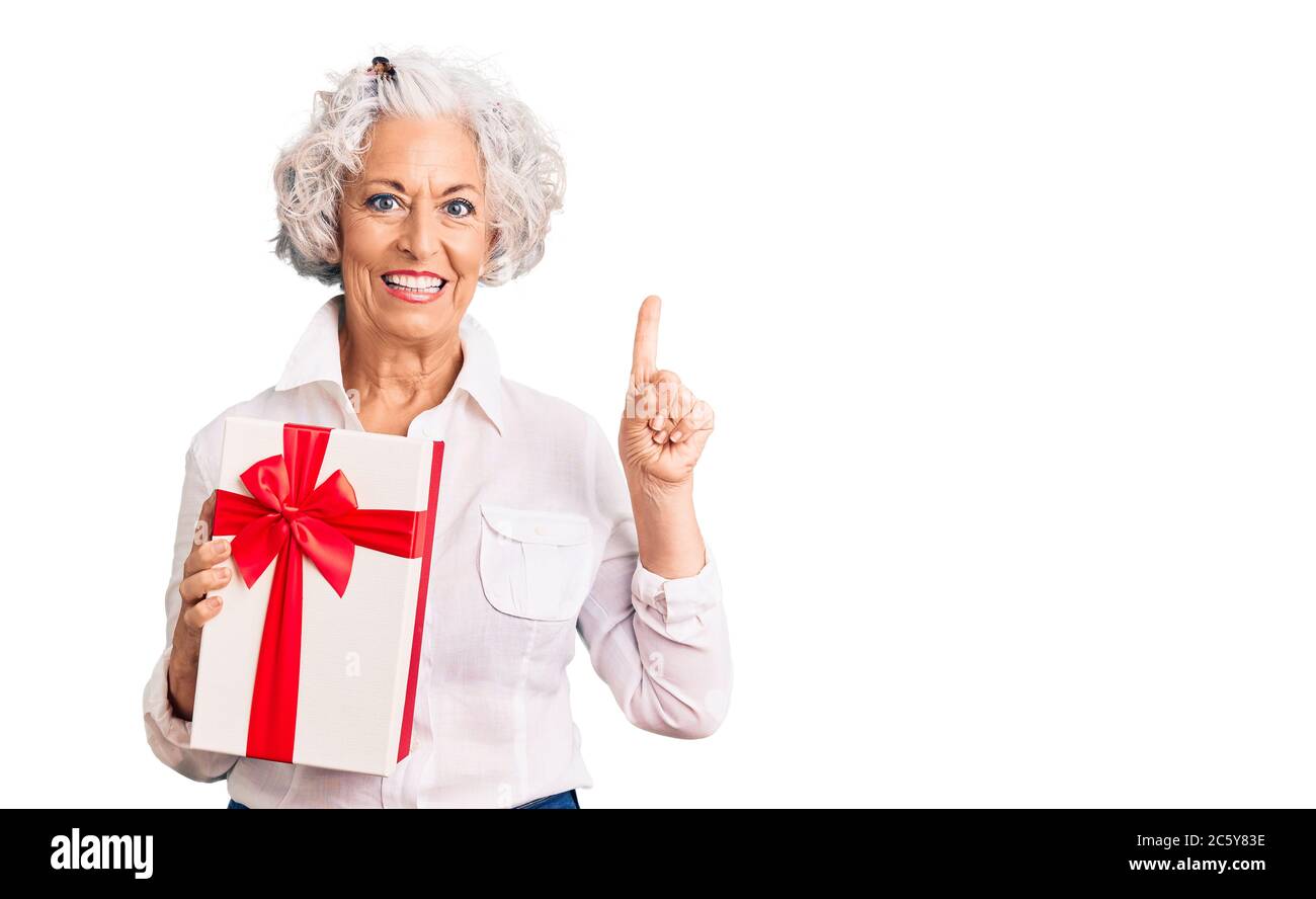 Senior grey-haired woman holding gift surprised with an idea or question pointing finger with happy face, number one Stock Photo