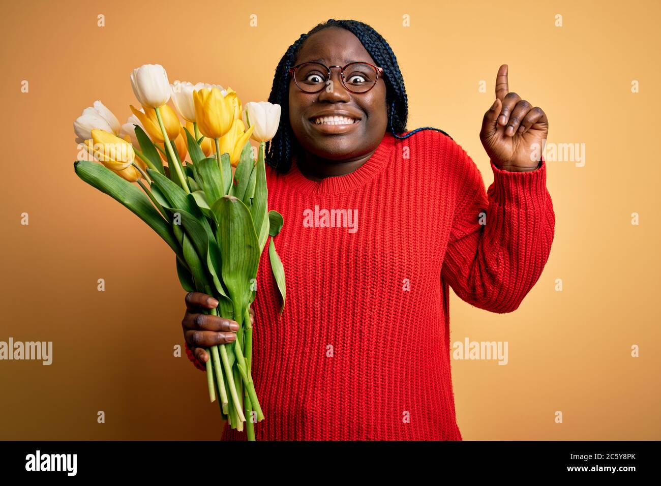 Young african american plus size woman with braids holding bouquet of yellow tulips flower smiling amazed and surprised and pointing up with fingers a Stock Photo