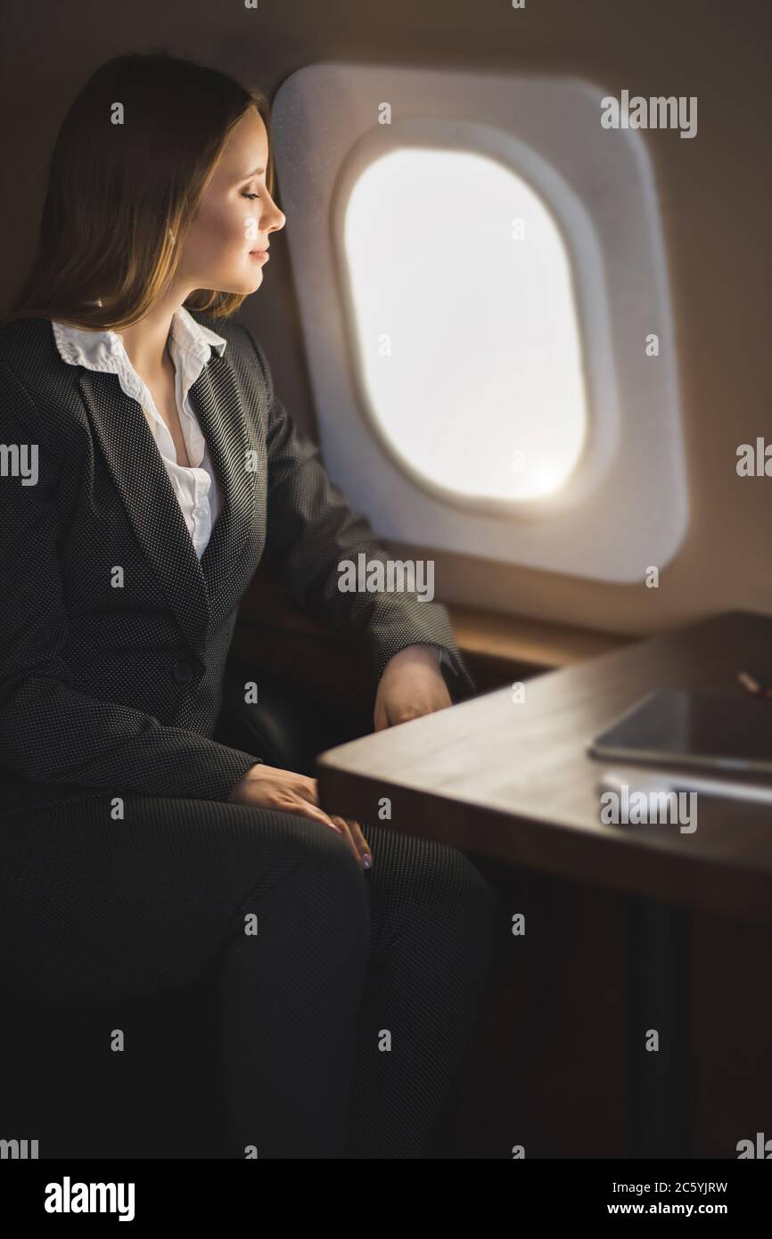 Happy attractive caucasian young businesswoman listening music using wireless earphones, looking at airplane window, admiring beauty of sky, enjoying Stock Photo