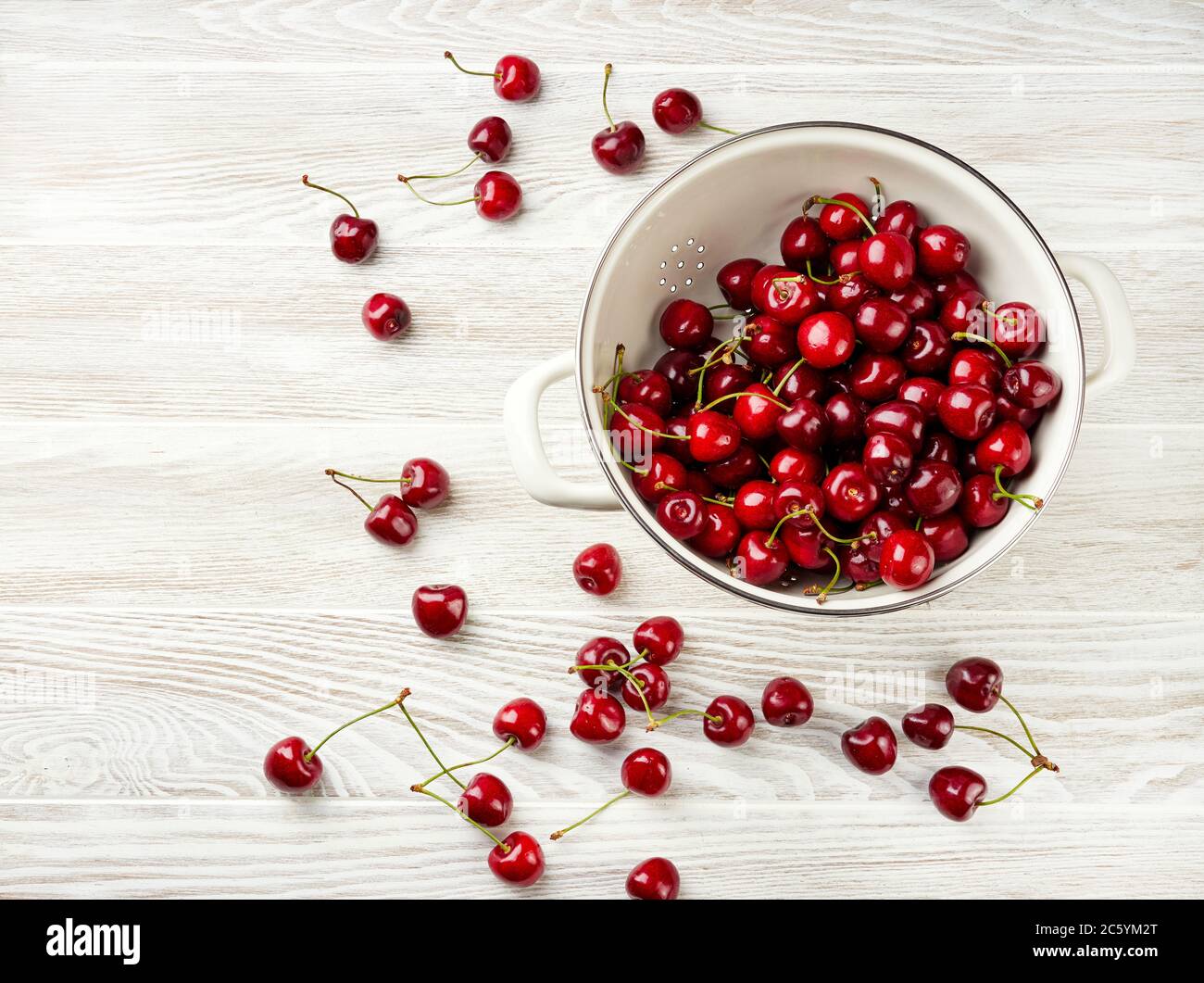 White strainer with fresh cherries on a wooden white background. Top view of colander with cherry berries. Stock Photo