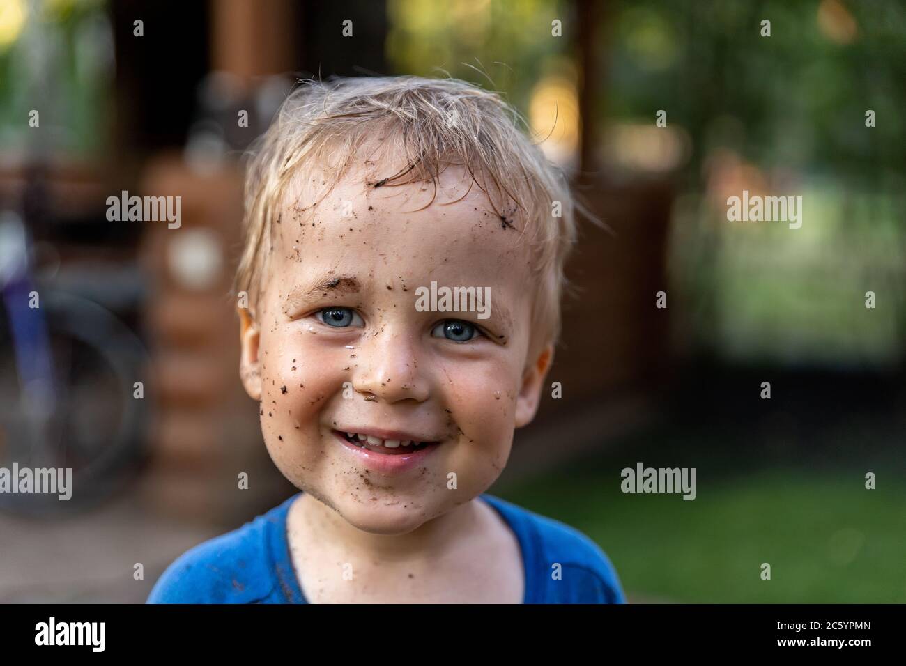 Cute adorable blond caucasian little happy toddler boy portrait with messy mus spots on face after playing watering garden at yard or countyside farm Stock Photo
