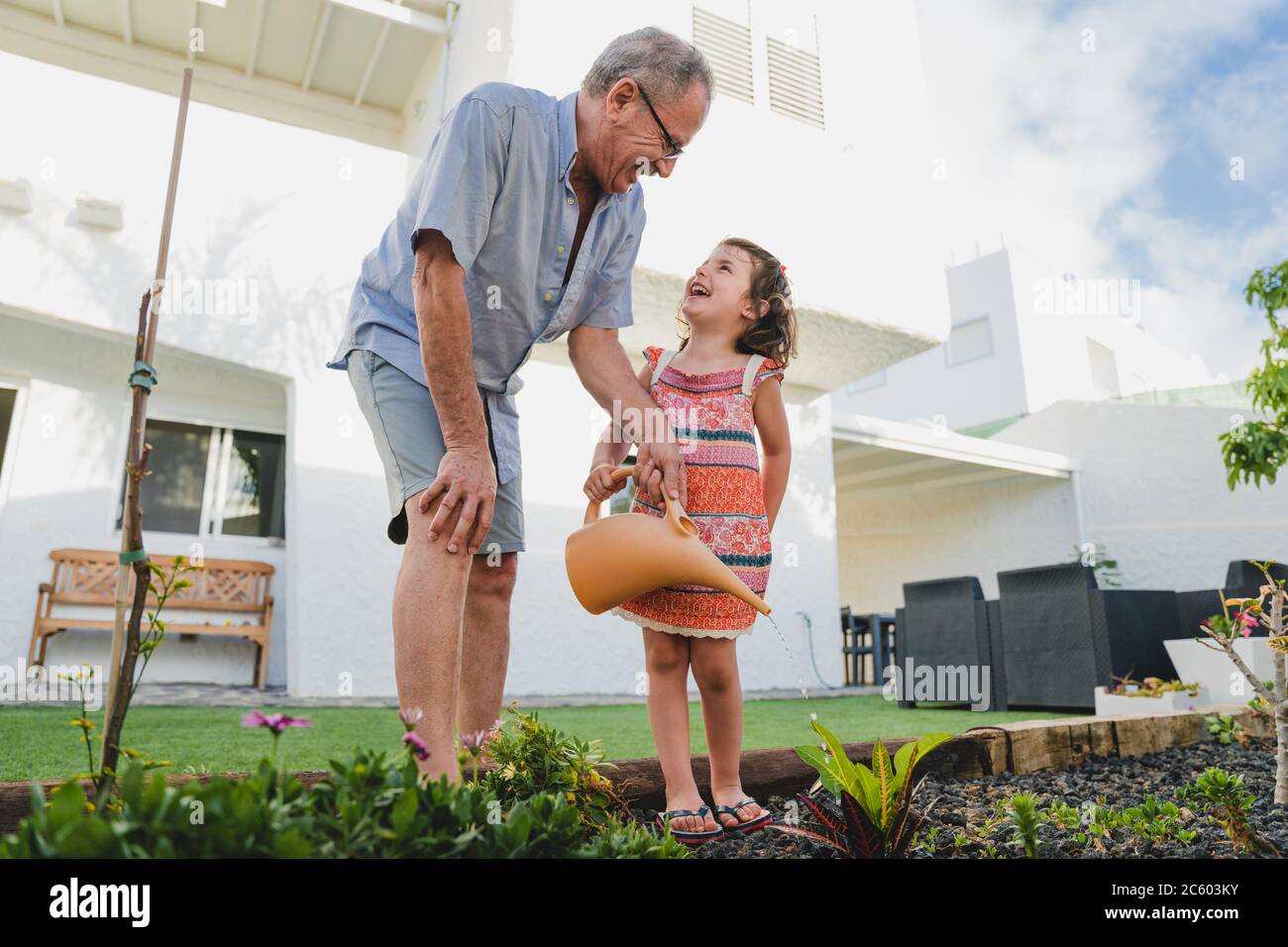 Smiling grandfather and granddaughter in backyard garden together, holding watering can have fun plants. Two generation fun time spend outdoor Stock Photo