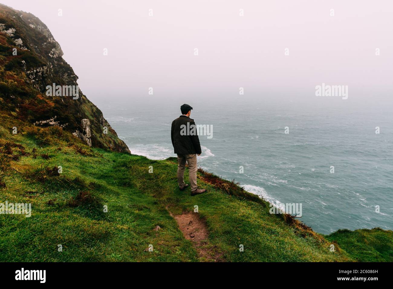 Man wearing cap stands alone in the cliffs of Brandon point in the Wild Atlantic Way of Ireland against the ocean a misty day. Stock Photo