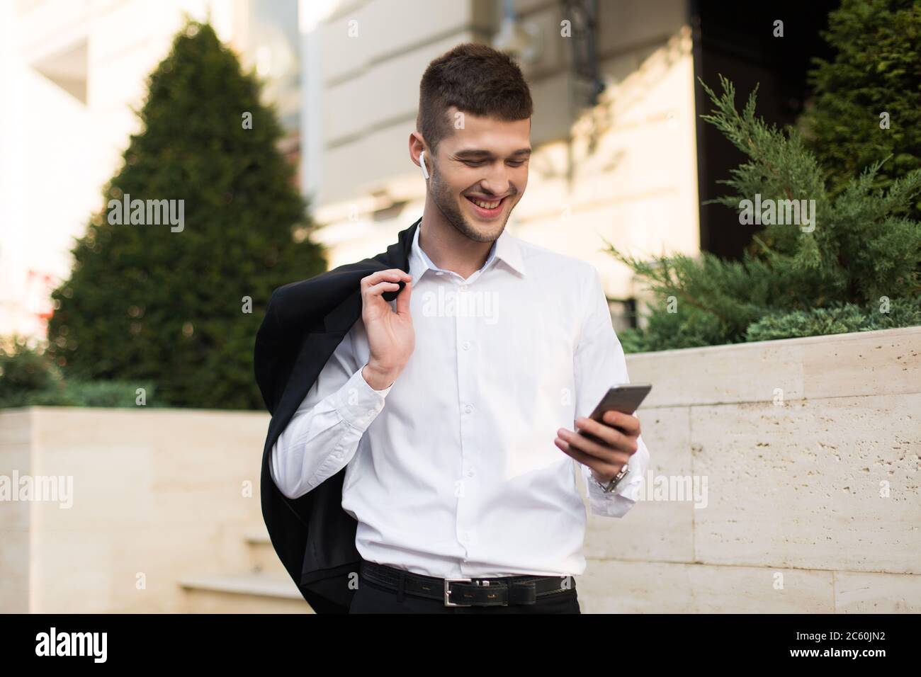 Young cheerful man in white shirt with wireless earphones holding black jacket on shoulder while happily using cellphone outdoor Stock Photo