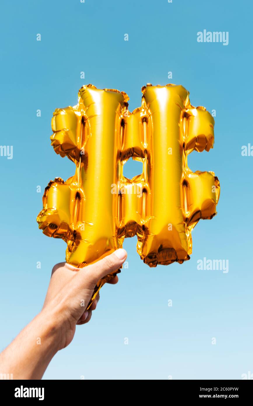 closeup of the hand of a young caucasian man holding a golden balloon in the shape of a hash symbol against the blue sky Stock Photo