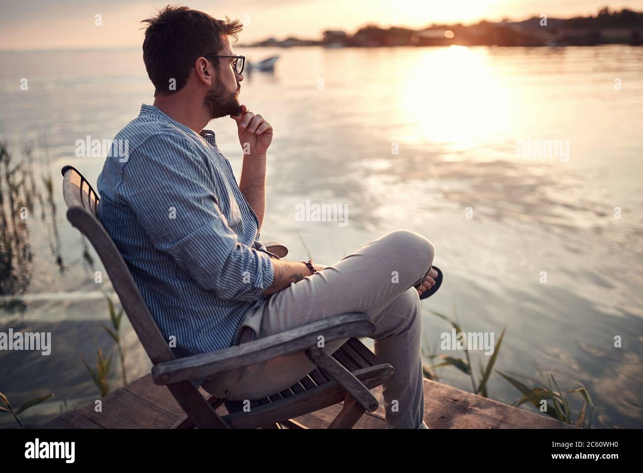 young beardy guy sitting alone on a river coast, enjoying the sunset, thinking Stock Photo