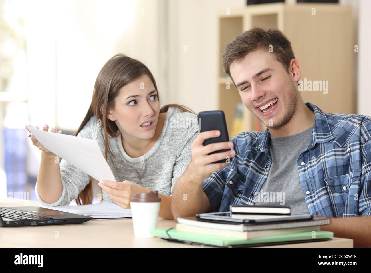 Angry student girl complaining showing homework to slacker classmate boy checking smartphone sitting on a desk at home Stock Photo