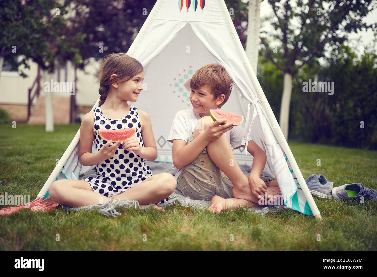 Summer time fun. Smiling boy and girl  at backyard in teepee and eats watermelon. Stock Photo