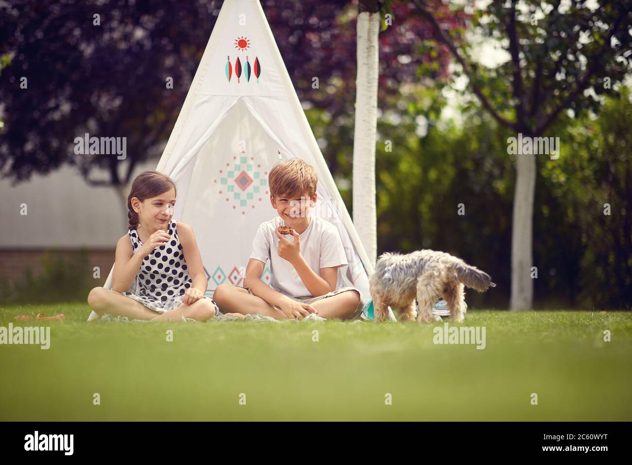 Summer time fun. Smiling children playing at backyard in teepee and eats cookies. Stock Photo