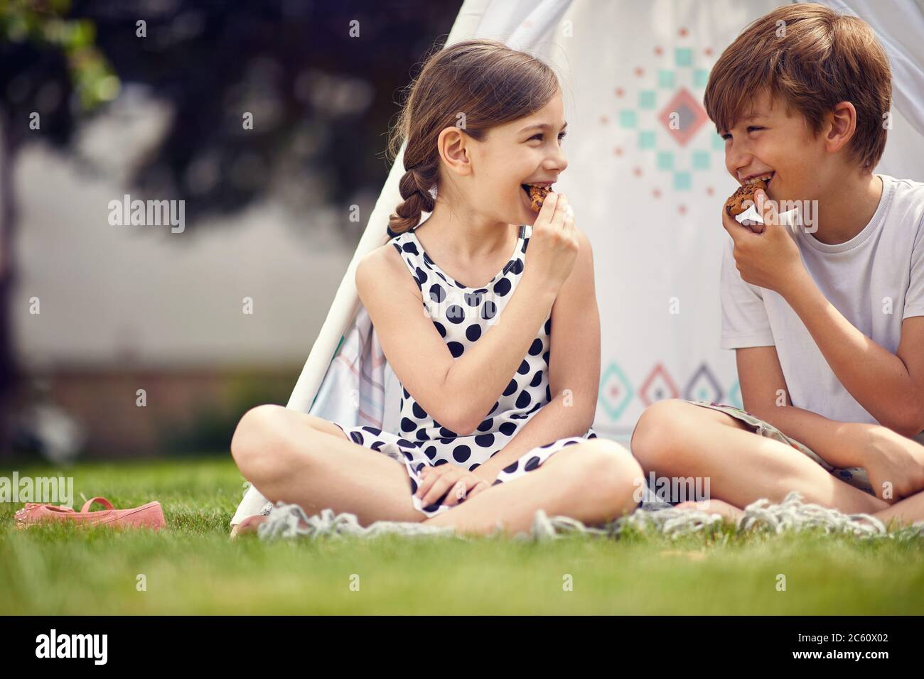 Summer time fun. Smiling kids playing at backyard in teepee and eats cookies. Stock Photo