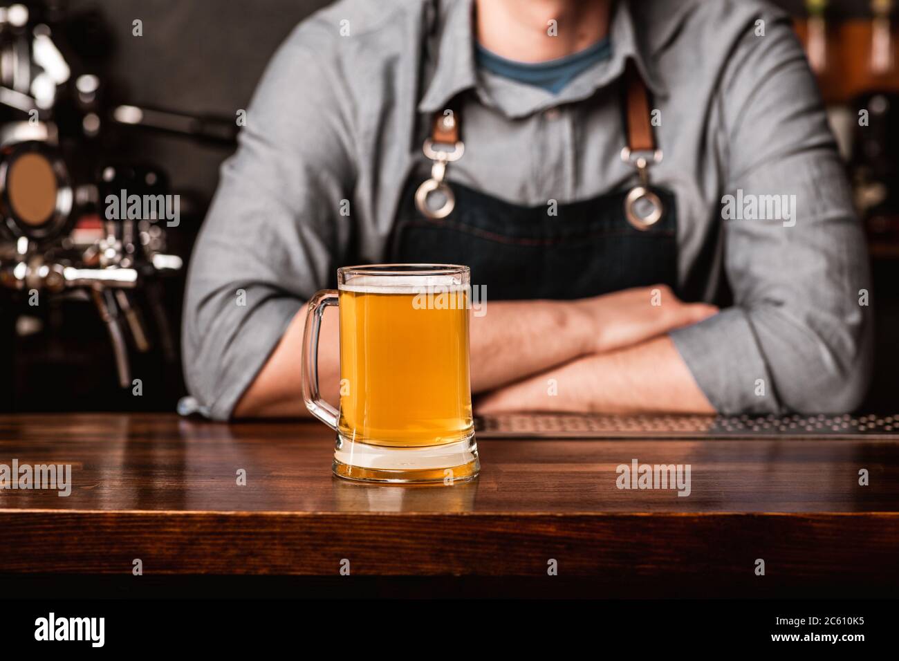 Barman in apron with crossed arms at bar and meets client with glass of light beer in interior of pub Stock Photo