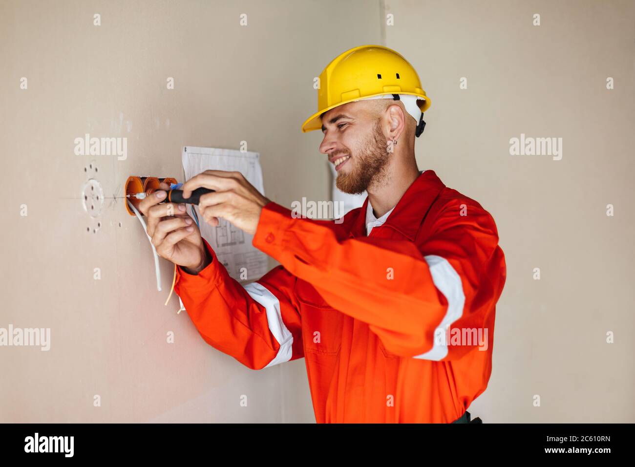 Smiling foreman in orange work clothes and yellow hardhat using Stock Photo