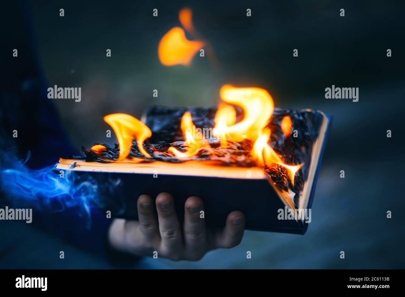 A man holds in his hand an old open book, the pages of which burn with a bright flame and emit blue magic smoke in the darkness. Fiery. Dangerous. Stock Photo