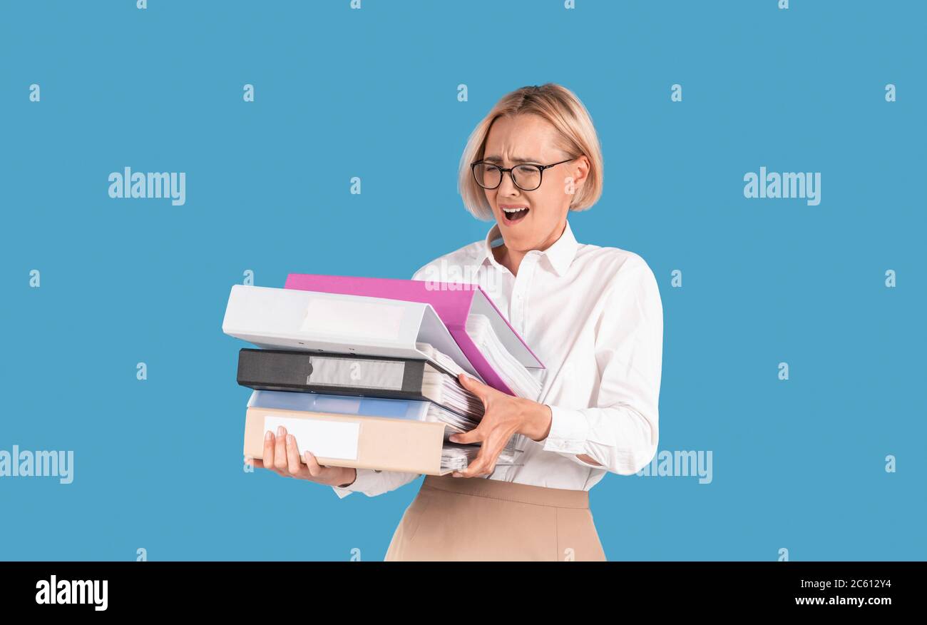 Stressed female secretary overloaded with paperwork, holding folders with documents on blue background Stock Photo