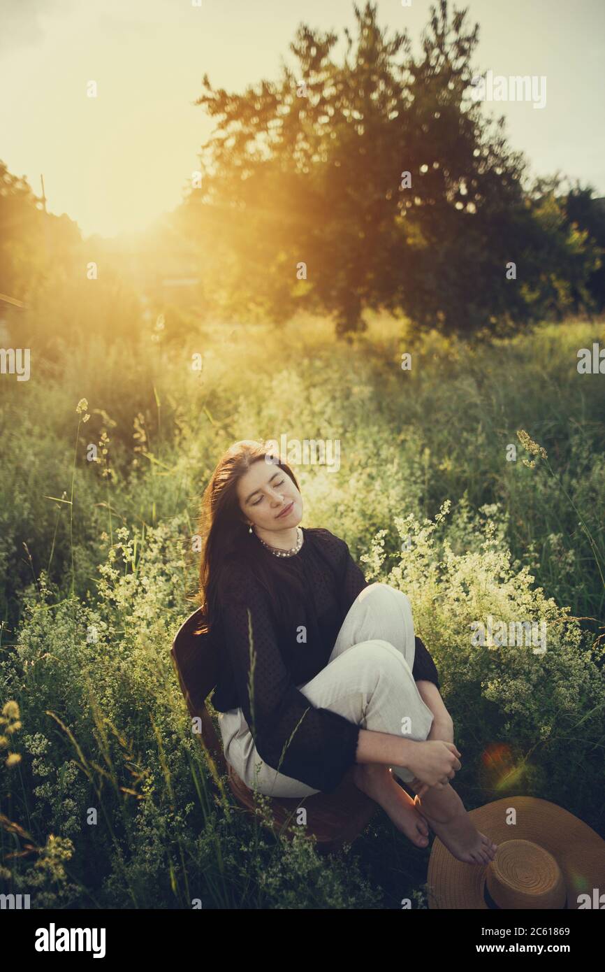 Slow living. Fashionable woman relaxing in summer countryside. Stylish elegant girl sitting on rustic chair in sunset light in summer meadow. Tranquil Stock Photo