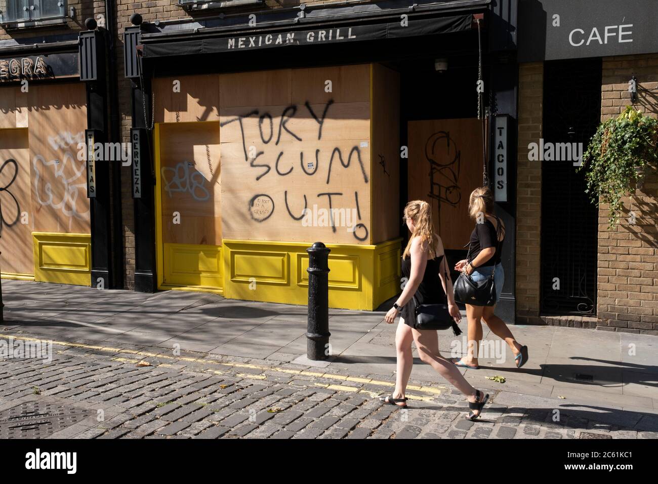 As the Coronavirus lockdown measures are set to ease further, and the quiet city starts coming to an end, some anti-Conservative party graffiti reading Tory Scum Out! on a boarded up restaurant in Soho on 25th June 2020 in London, England, United Kingdom. As the July deadline approaces and government will relax its lockdown rules further, the West End remains quiet, while some non-essential shops are allowed to open with individual shops setting up social distancing systems. Stock Photo