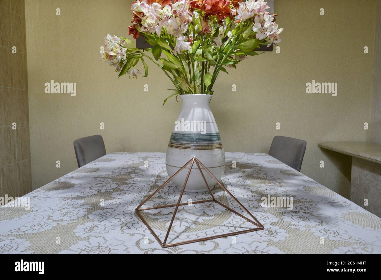 Copper pyramid, with Astromelia vase, Alstroemeria Hybrida on a travertine marble table base with white lace tablecloth, with chairs and white wall in Stock Photo