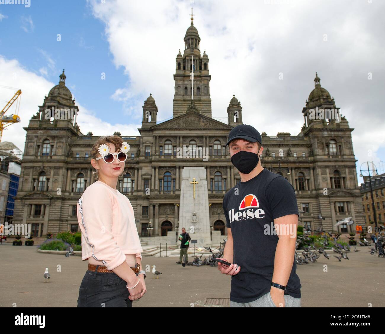 Glasgow, Scotland, UK. 6th July, 2020. Pictured: A couple seen rocking their summer attire in George Square with a lady wearing daisy pattern sun glasses and the gentleman wearing a black face mask and black baseball cap, with the City Chambers as a backdrop. People in George Square. Bars and Restaurants re-open in Glasgow as lockdown restrictions ease. From today Pubs, bars, cafes and restaurants in England, Scotland and Northern Ireland are welcoming customers for the first time since lockdown began in March. Credit: Colin Fisher/Alamy Live News Stock Photo