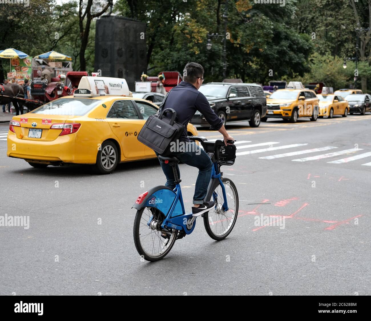 Adult member of the public seen riding a bicycle beside a moving New York taxi past the popular tourist attraction of Central Park in Manhattan. Stock Photo