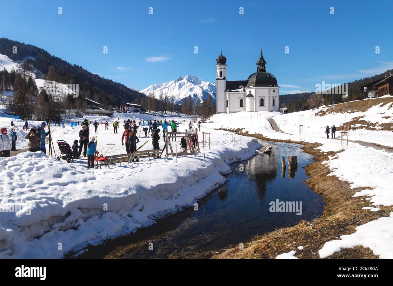 Seefeld, Tirol, Austria - 8 March 2020: Cross-country skiers on sunny track along a river and a pitoresque church reflecting in the water Stock Photo