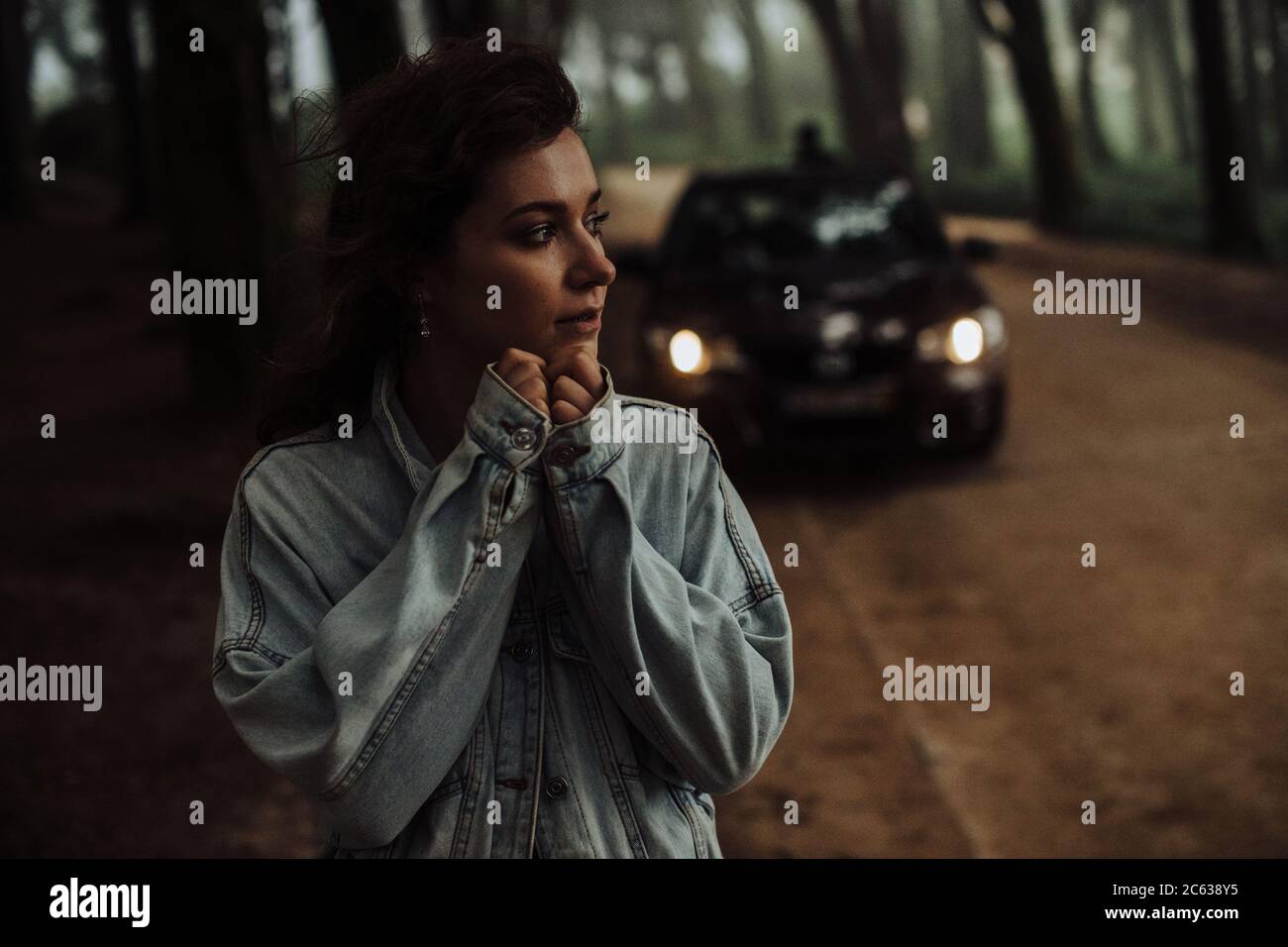 Close up of young woman standing alone in forest with car behind Stock Photo