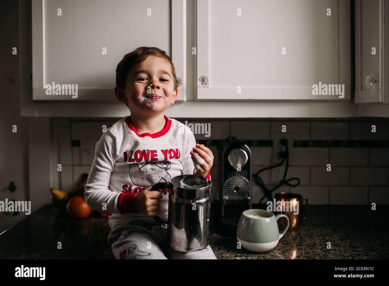 Young Boy Sitting On Kitchen Counter With Frothed Milk On Face Stock