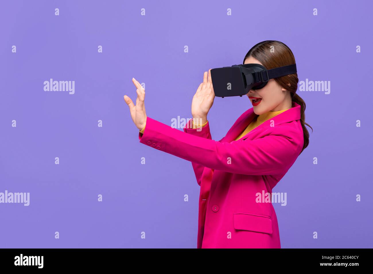 Young woman wearing virtual reality or VR glasses reaching hand out to touch something on purple isolated background Stock Photo