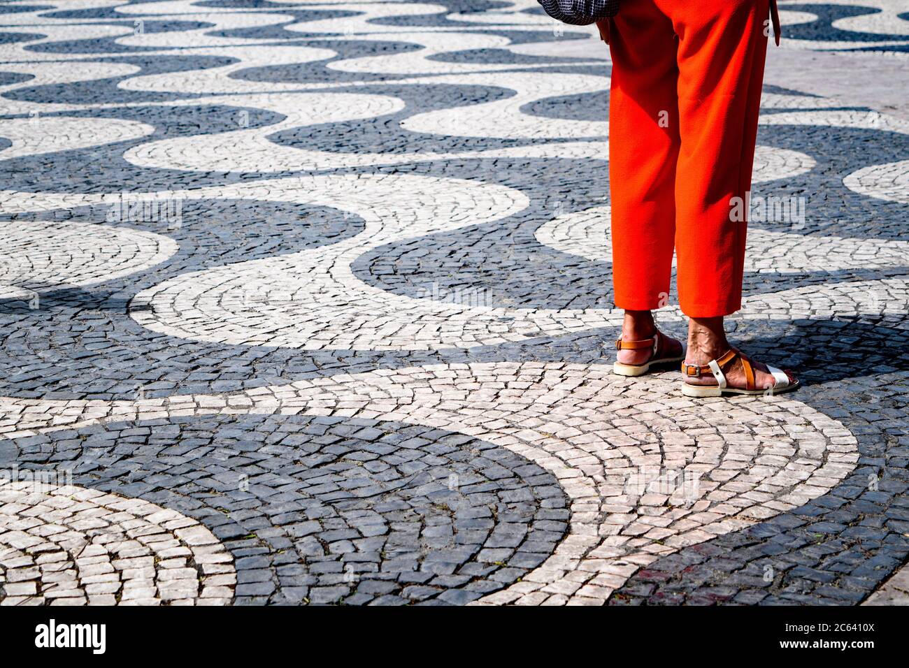 A woman's orange pants create a vivid pop of color against the black and white wave-patterned paving stones in Lisbon's Rossio Square. Stock Photo