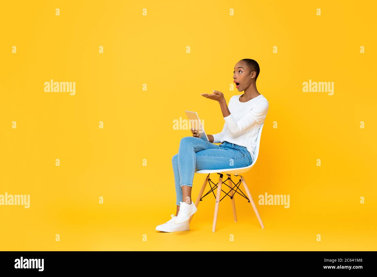 Young surprised African American woman sitting and opening hand looking up to copy space while using tablet computer on isolated yellow background Stock Photo