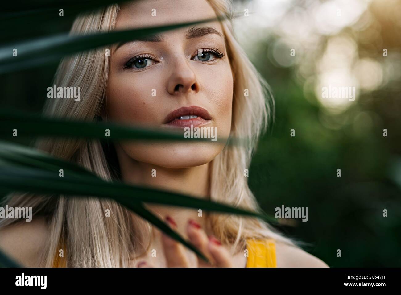 Closeup beauty portrait of lovely caucasian blonde haired woman with clean skin, natural makeup, beautiful eyes, standing behind tropical leaf. Vertic Stock Photo