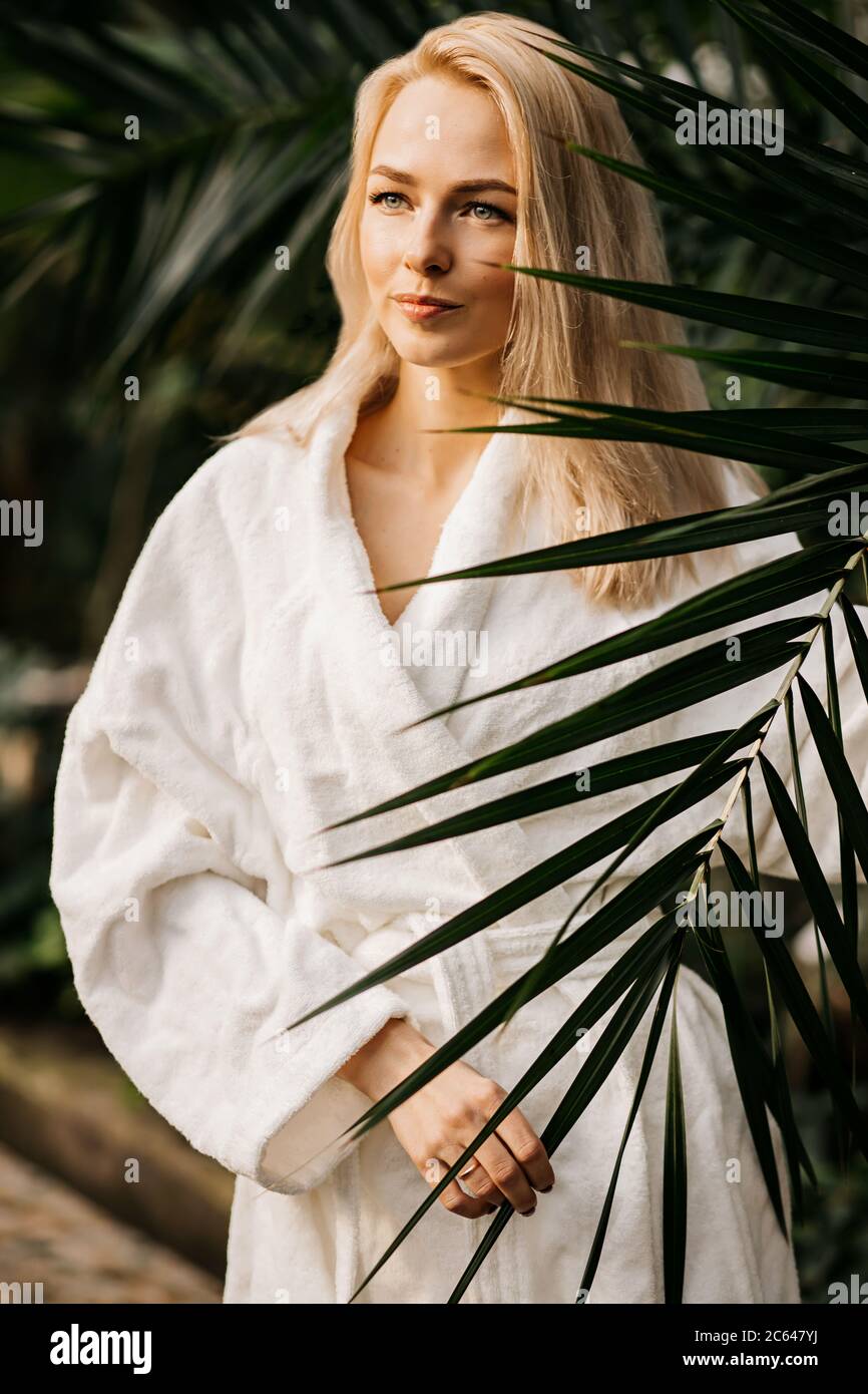 Closeup a beautiful portrait of a young Spa woman in a white coat against the background of an exotic forest with palm leaves on her hands and a smile Stock Photo