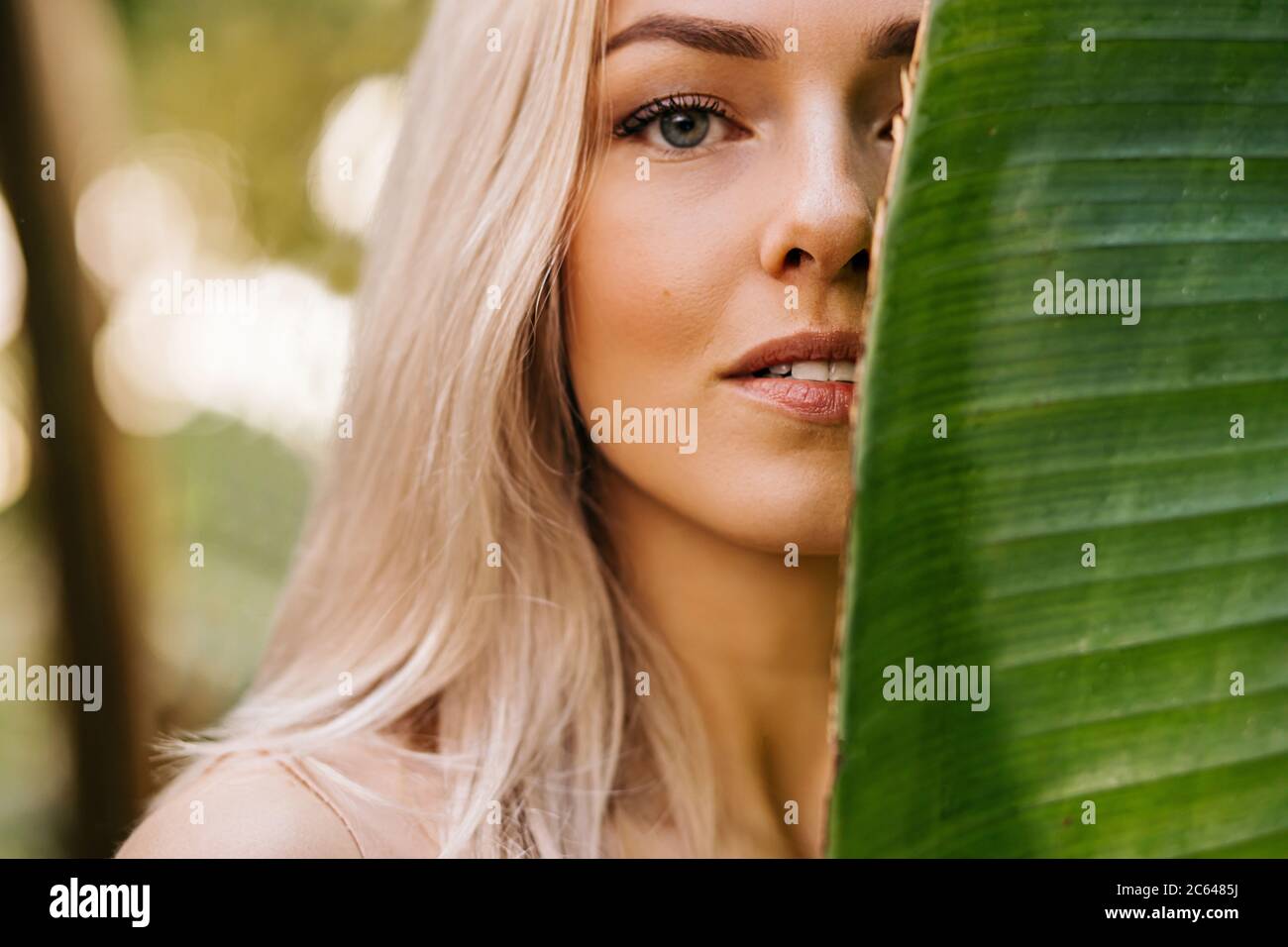 Closeup portrait of pretty caucasian lady with blonde hair, natural makeup, clean fresh skin, hiding half of face behind big tropical leaf. Skin care, Stock Photo