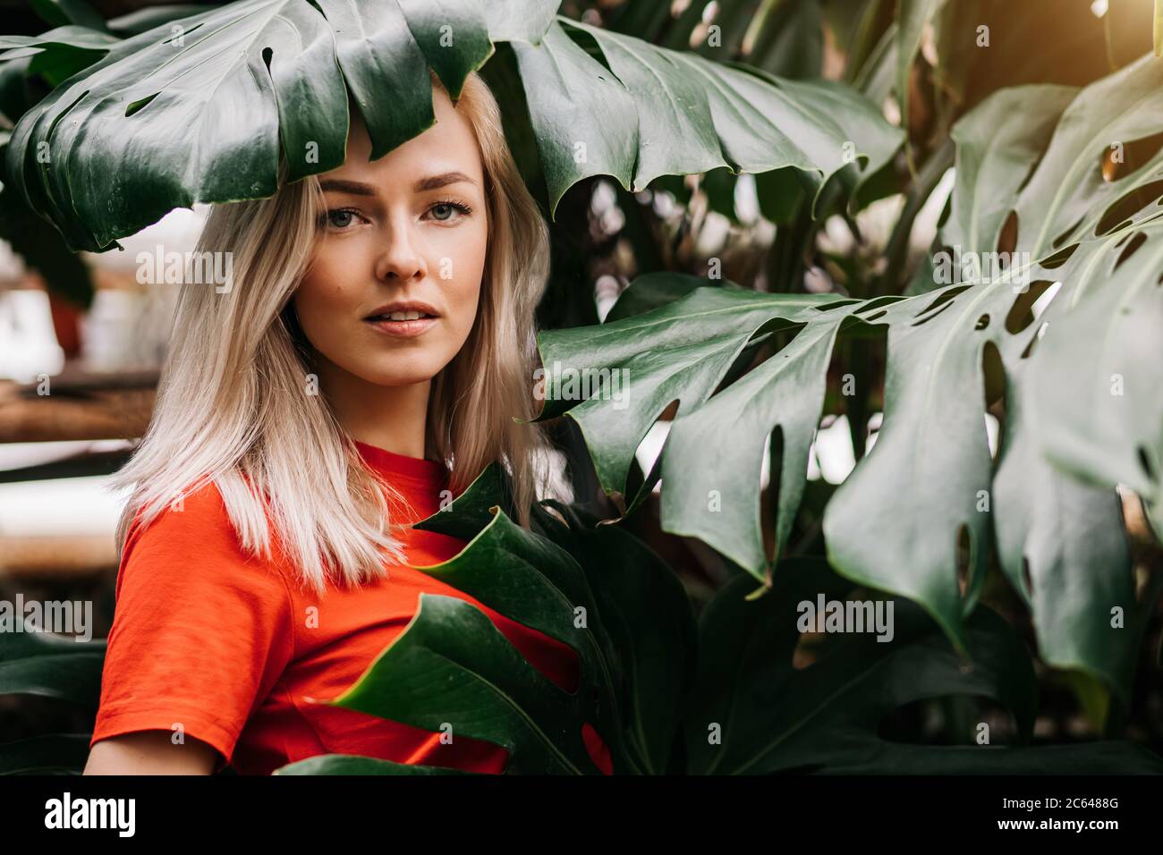 Blonde lady with natural makeup posing in rainforest. Gorgeous caucasian woman in red t shirt standing among monstera leaves, enjoying summer vacation Stock Photo