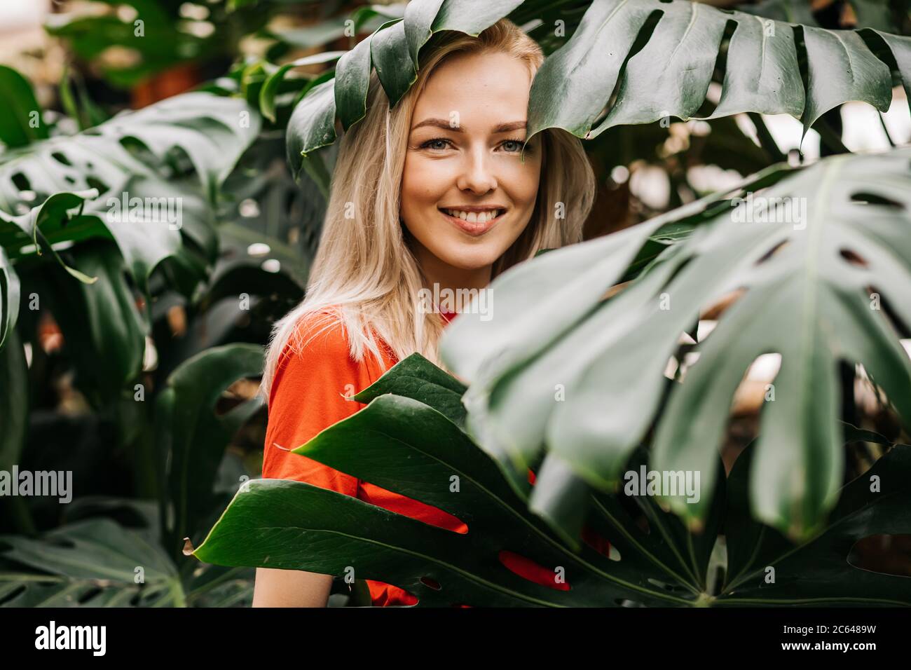 Cheerful charming caucasian blonde woman wearing red t shirt enjoying time in forest, smiling with white teeth standing among tropical leaves. Nature Stock Photo