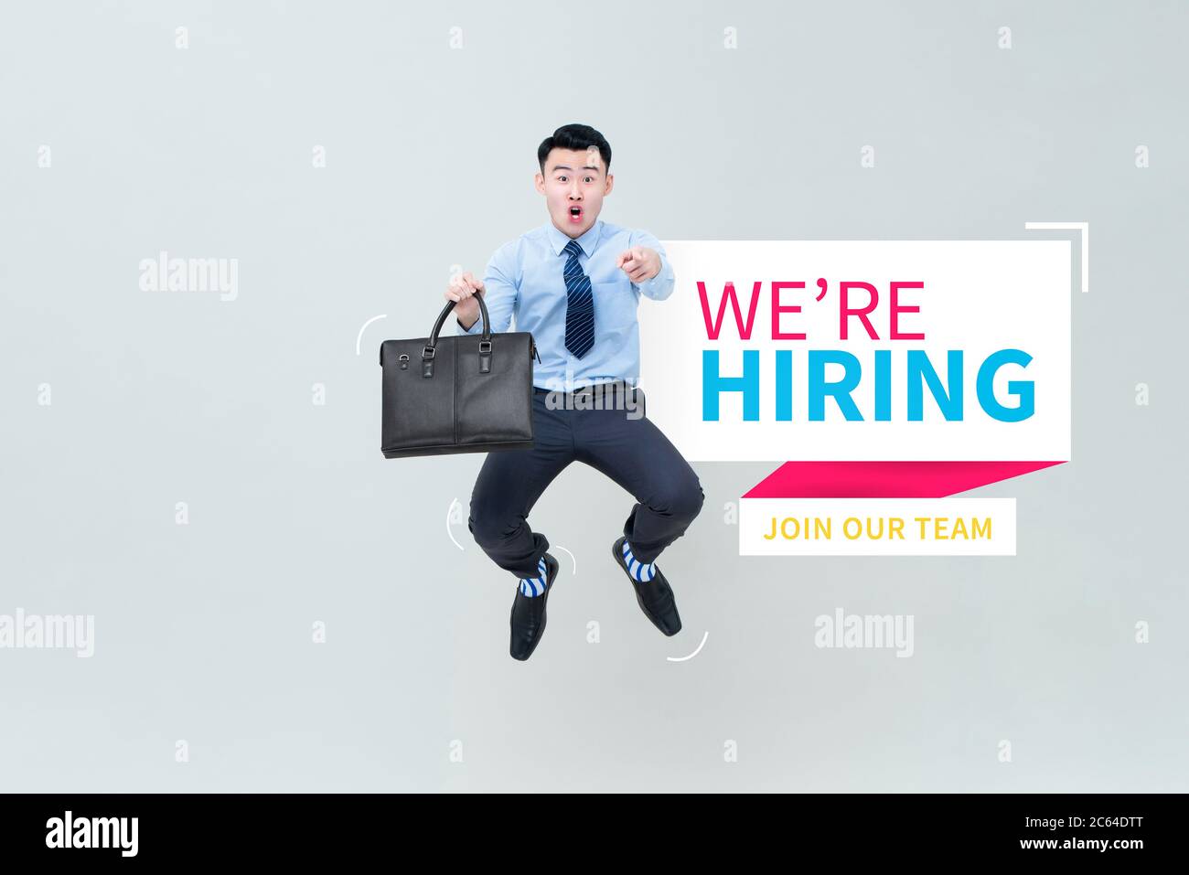 Excited young Asian businessman jumping and pointing finger at camera next to We're hiring and join our team text on light gray background Stock Photo