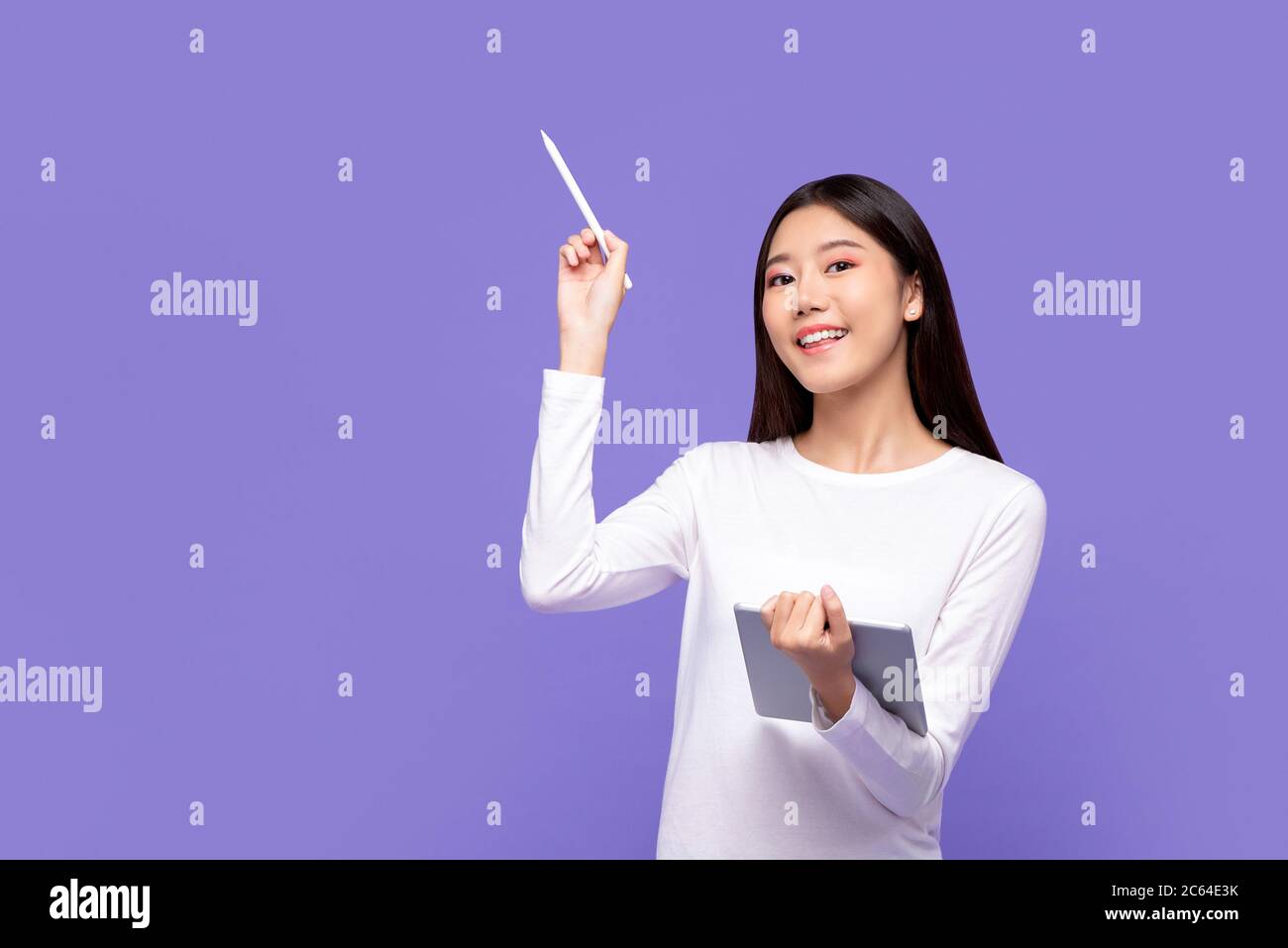 Smiling beautiful Asian woman in white attire pointing with stylus to copy space and holding digital tablet isolated on purple background Stock Photo