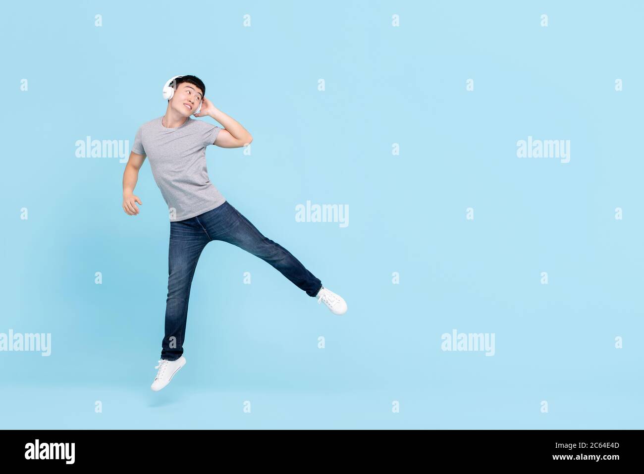 Smiling energetic young Asian man wearing headphones listening to music jumping and looking at copy space on light blue isolated background Stock Photo