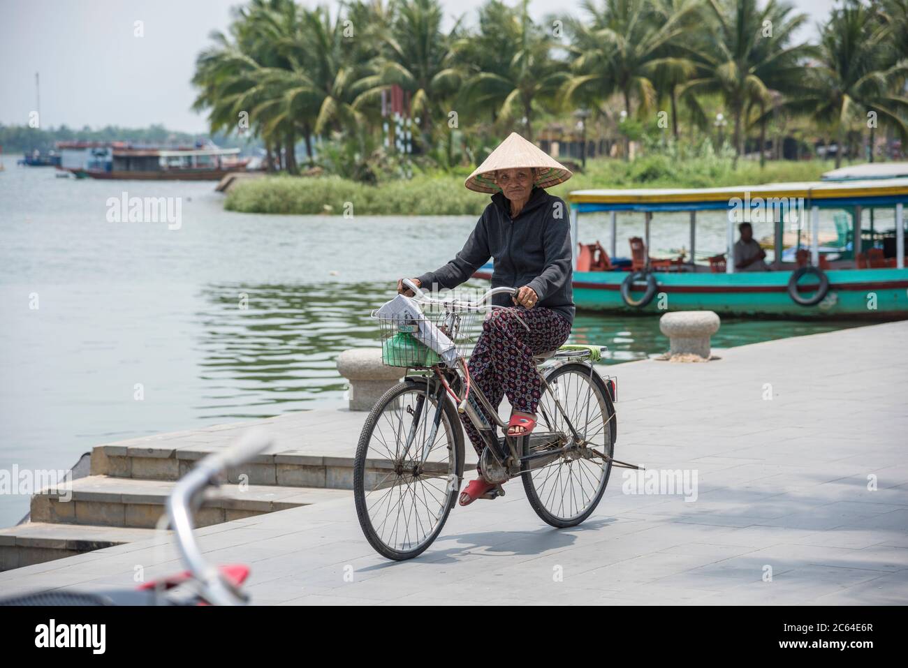 Elderly Vietnamese lady riding a biycle along the river frontage in Hoi An  Vietnam. Stock Photo