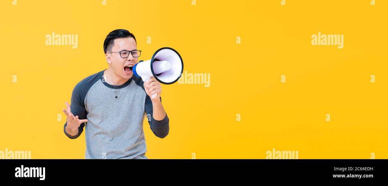Angry young Asian man shouting into megaphone isolated on yellow banner background with copy space Stock Photo