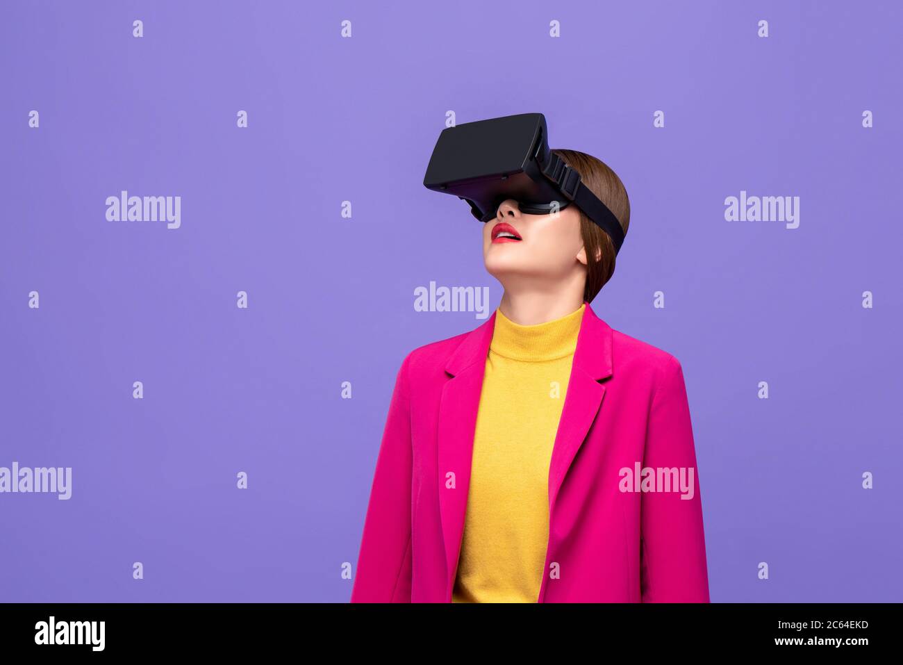 Woman in colorful attire raising head and watching 3D simulation video from virtual reality or VR glasses on purple background Stock Photo