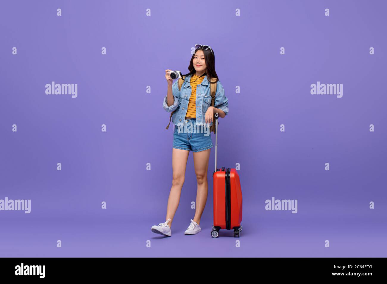 Young smiling Asian tourist girl in casual attire with camera and baggage isolated on colorful purple background Stock Photo