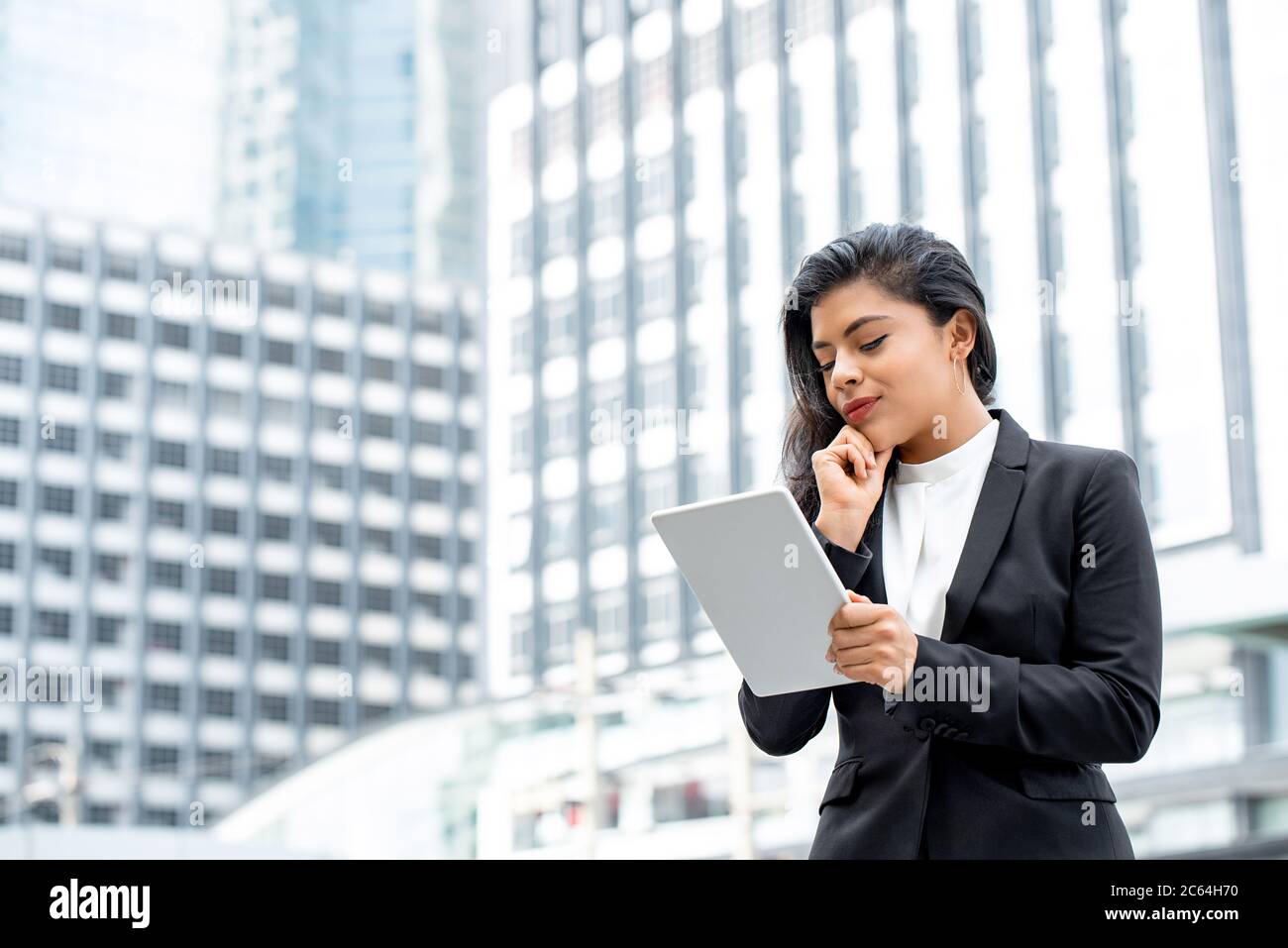 Portrait of smiling young confident Hispanic businesswoman looking at tablet in urban city background Stock Photo