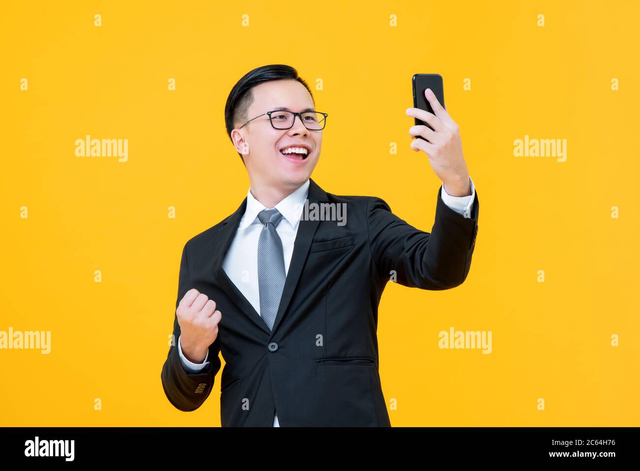 Portrait of happy confident Asian businessman taking a selfie doing a success closed fist gesture in isolated studio yellow background Stock Photo