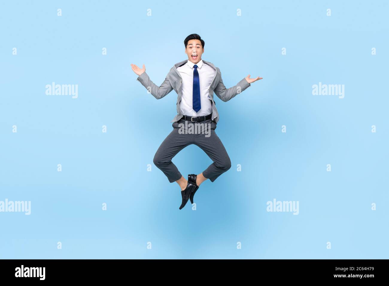 Full length playful portrait of happy ecstatic young Asian businessman jumping in mid-air doing wacky fun gesture in isolated studio blue background Stock Photo