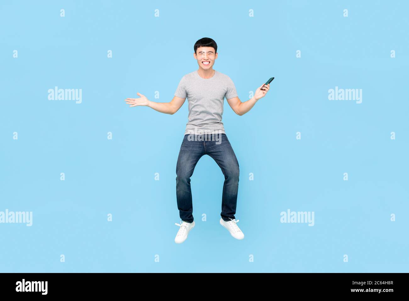 Fun full length portrait of happy smiling Asian man jumping in mid-air while holding smartphone in isolated studio blue background Stock Photo