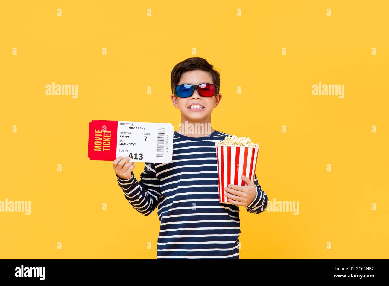 Fun portrait of happy young Asian boy wearing 3D glasses holding popcorn and movie ticket in isolated studio yellow background Stock Photo