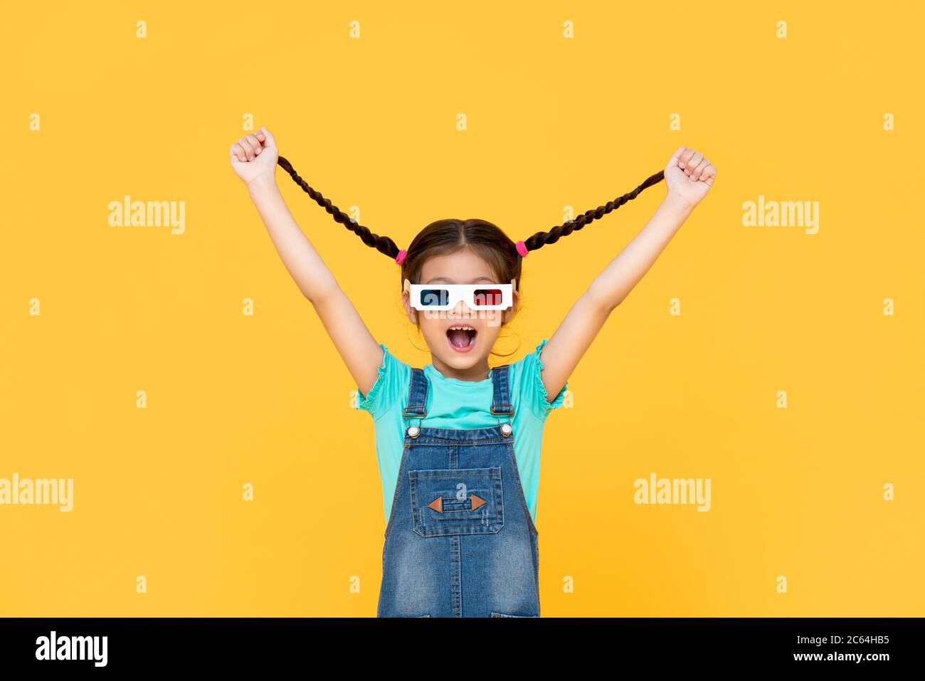 Fun Portrait of excited young girl wearing 3D cinema glasses with both arms raised pulling her hair in isolated studio yellow background Stock Photo