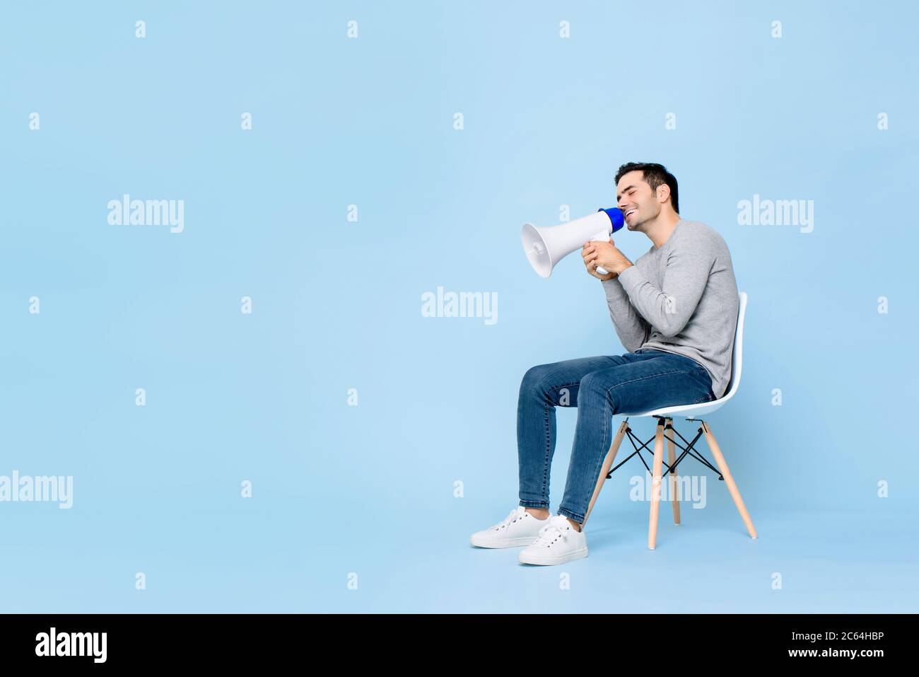 Portrait of smiling young handsome Caucasian man sitting while holding and announcing on megaphone in isolated studio blue background Stock Photo