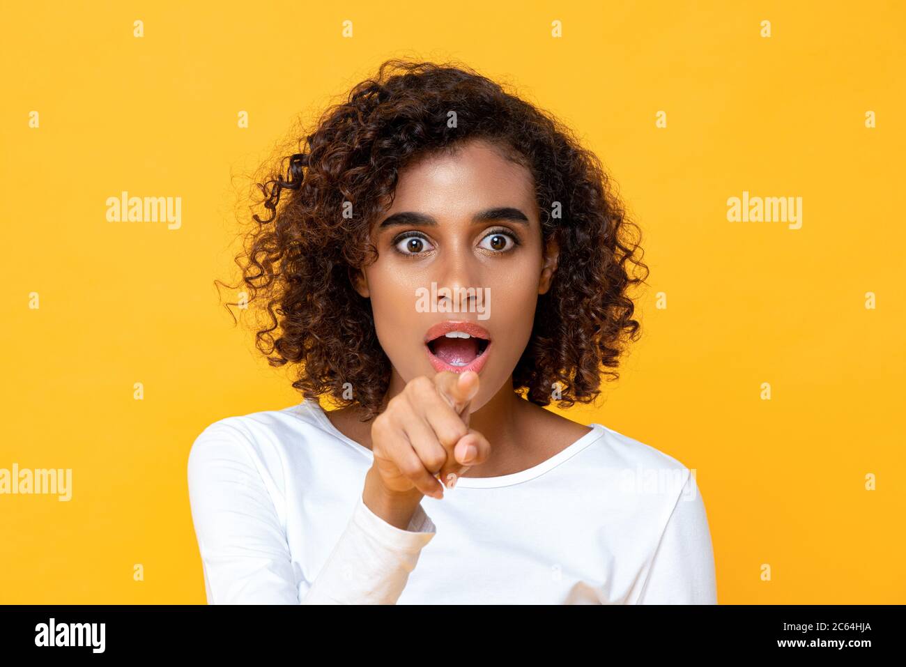 Close up portrait of attractive young African American woman looking shocked while pointing finger at front in isolated studio yellow background Stock Photo