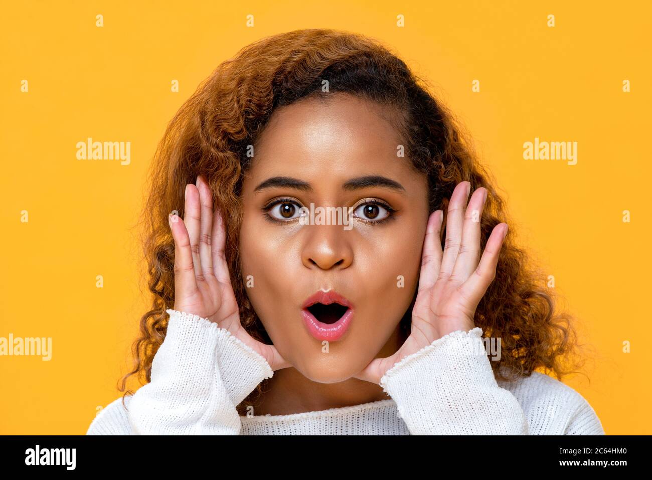 Fun close up portrait of surprised young beautiful African American woman  with hands cupped around opened mouth in isolated studio yellow background Stock Photo